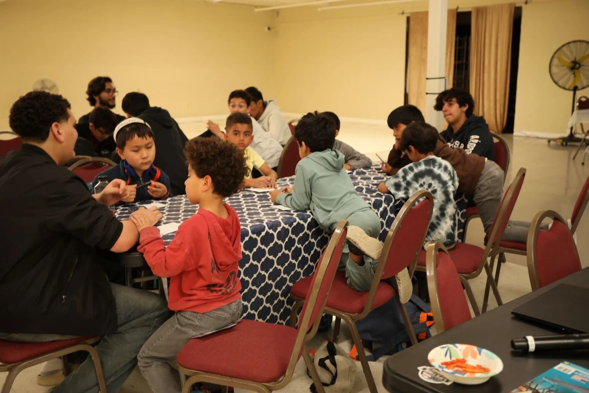 Children and teen mentors working on activities around tables with navy patterned tablecloths in a warm-lit hall.