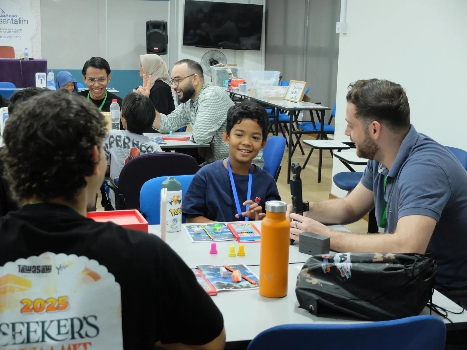 Smiling boy with a lanyard at a classroom table covered with activity materials, adult mentor beside him.