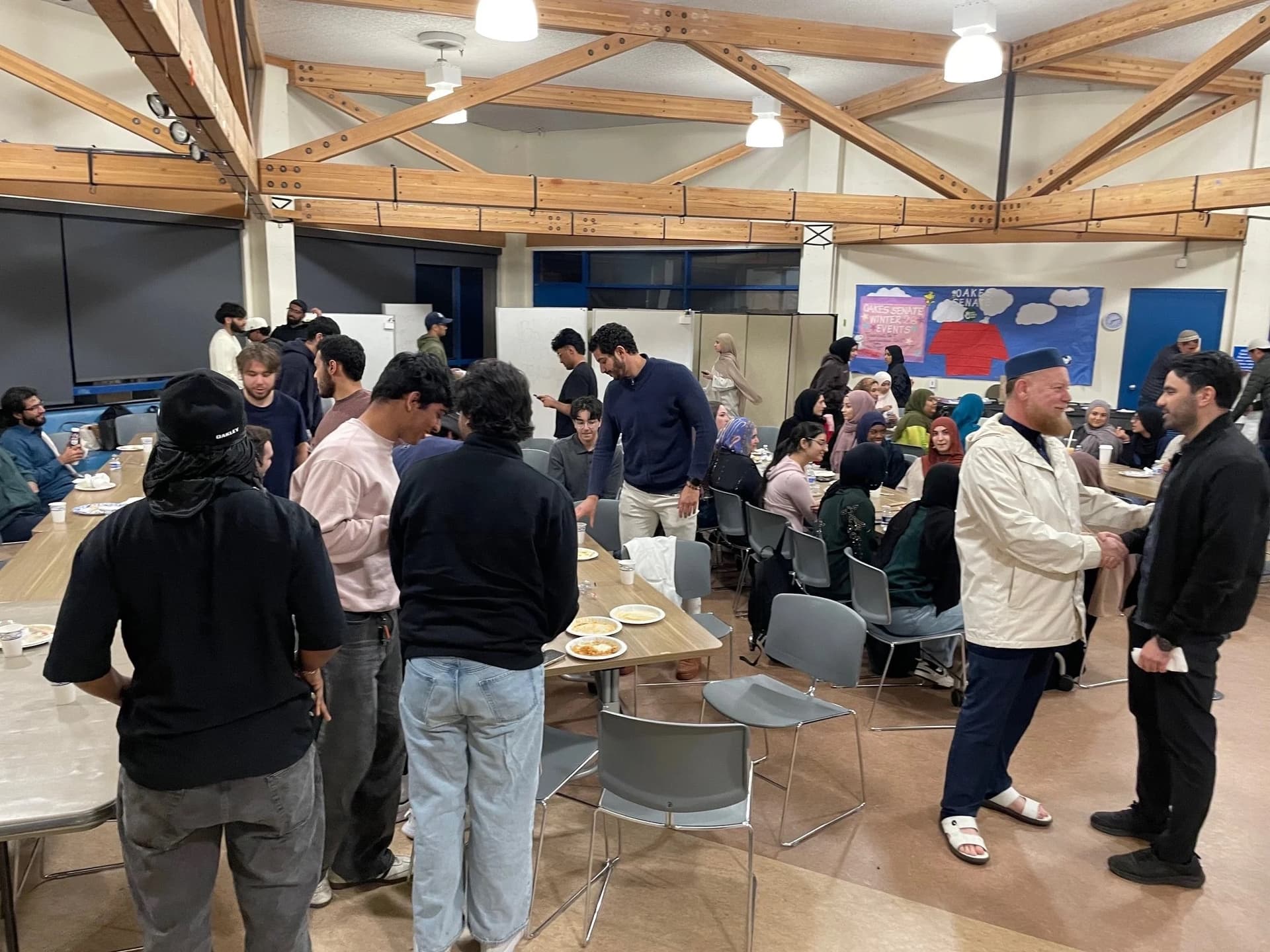 Young adults gathered around long tables for a shared meal in a hall with exposed timber ceiling beams.