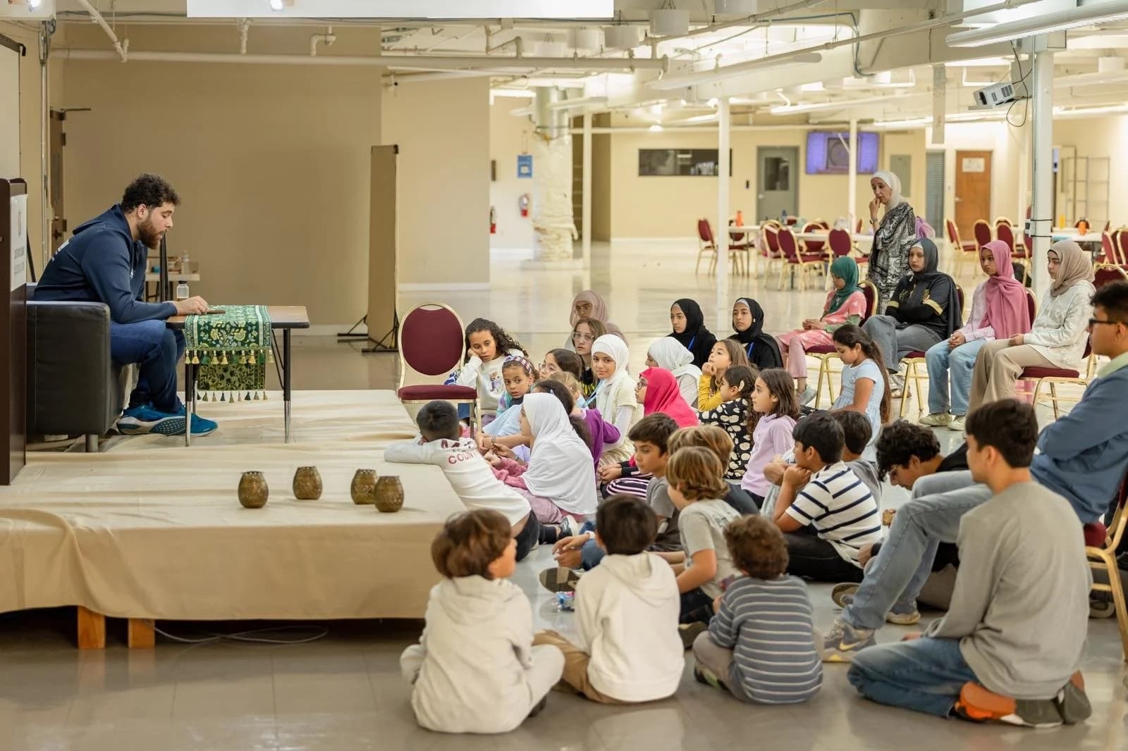 Children seated on the floor in a bright hall listening to an instructor seated at a low table.