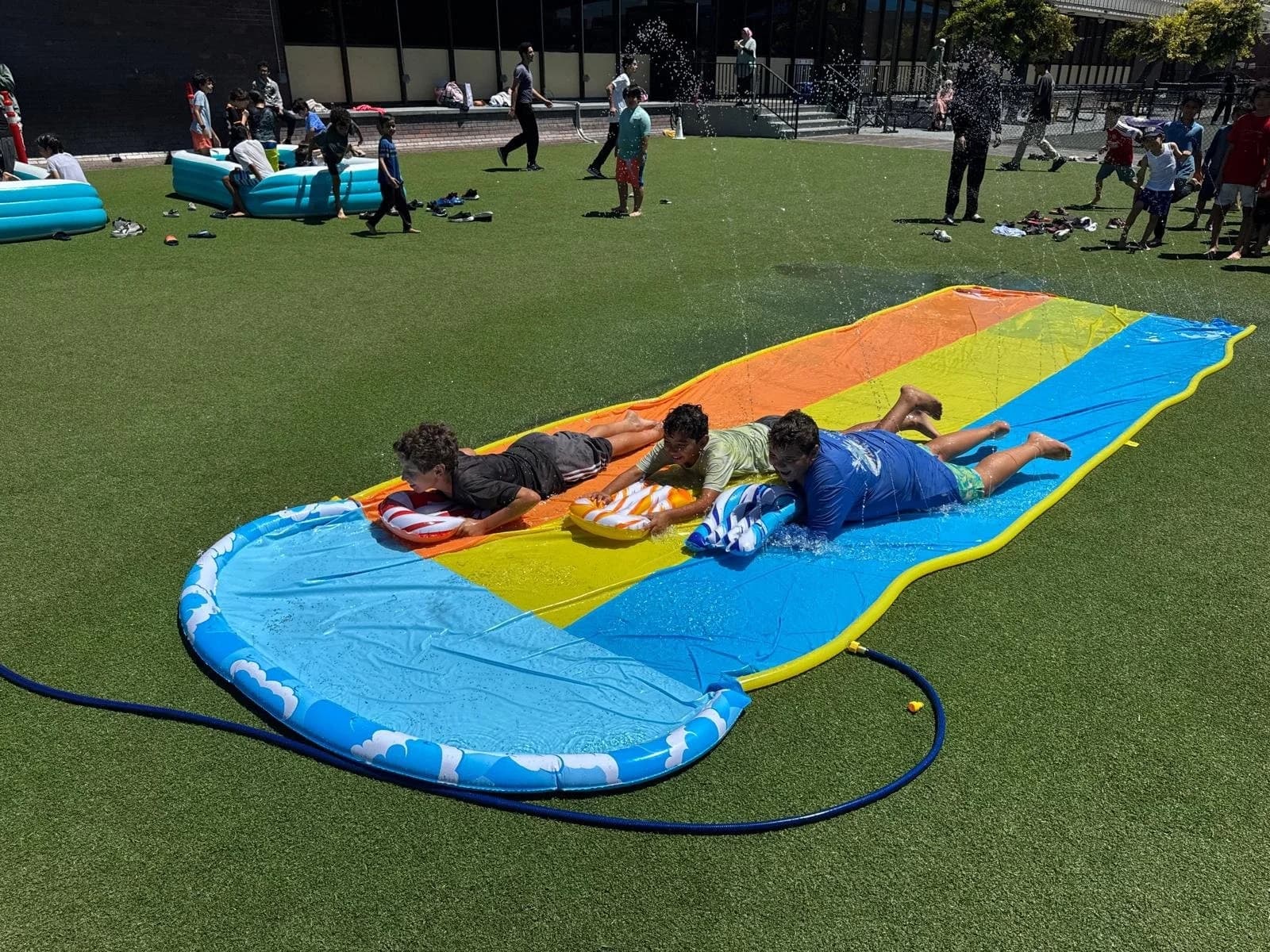 Three children sliding on a colourful inflatable water slide on artificial turf, others playing in the background.