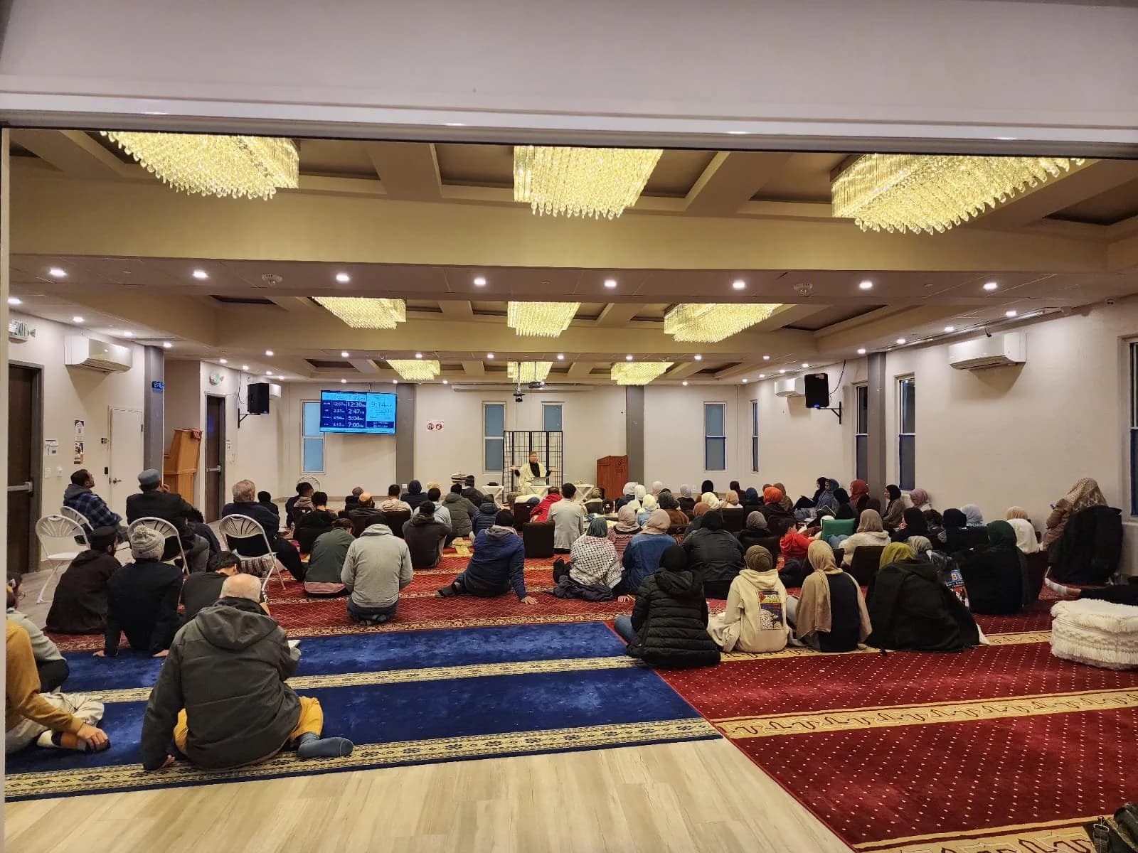 Audience seated on carpet facing a seated speaker in a hall with crystal chandeliers and a prayer-times screen.
