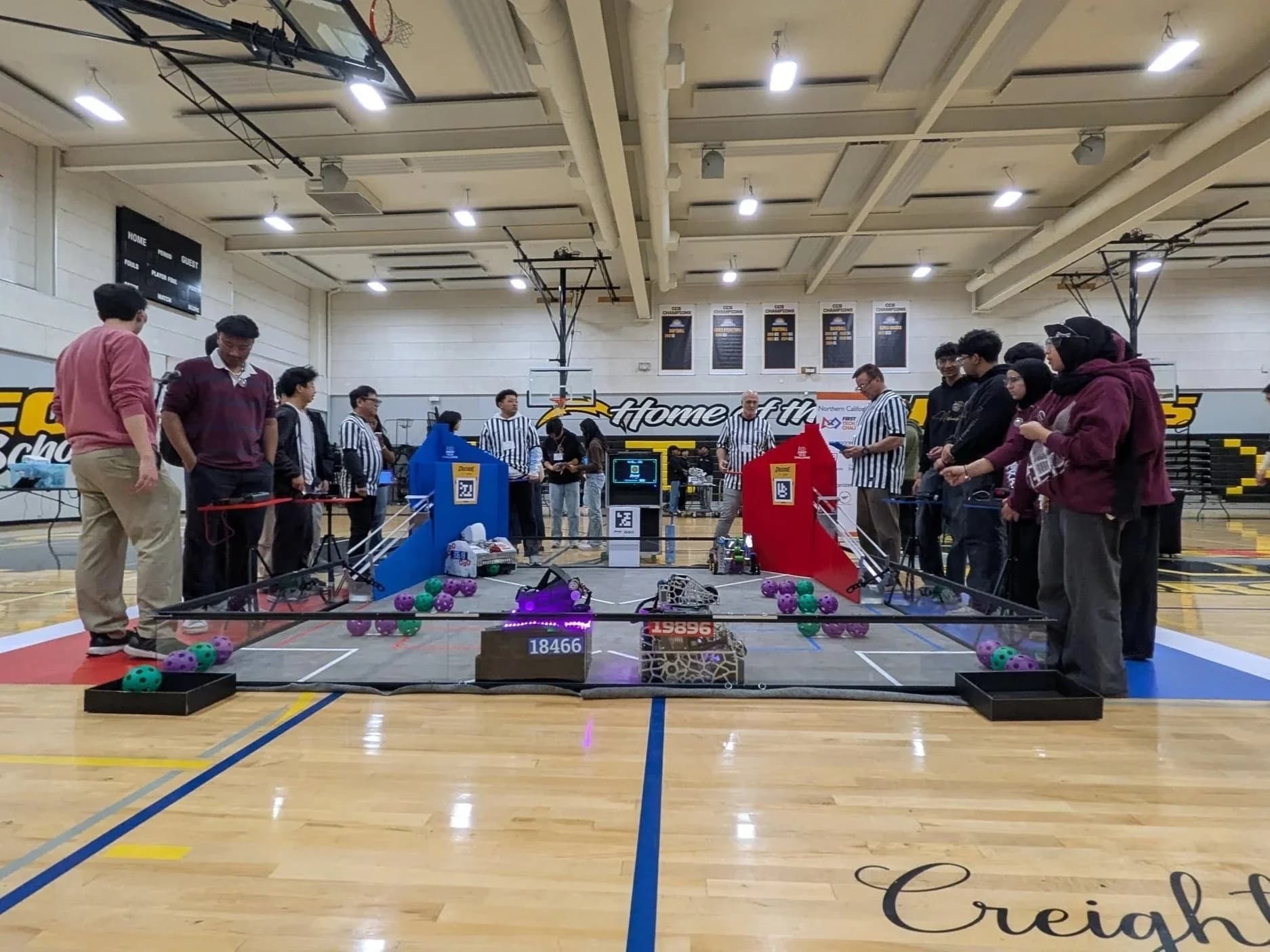 Robotics competition field in a school gym, with teams and referees in striped shirts standing around the arena.