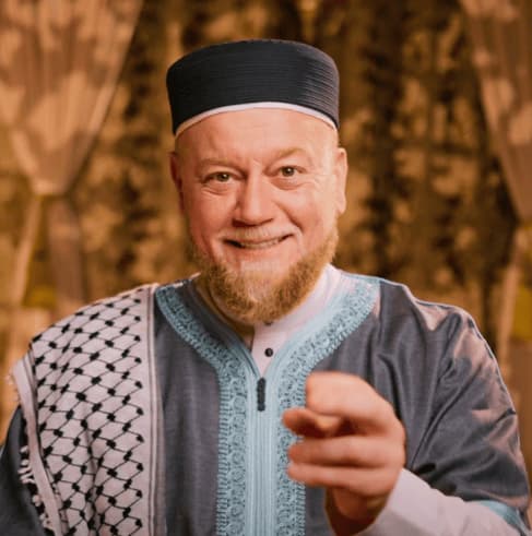 Shaykh Alauddin ElBakri, smiling, in a dark kufi and a blue thobe with a keffiyeh over one shoulder
