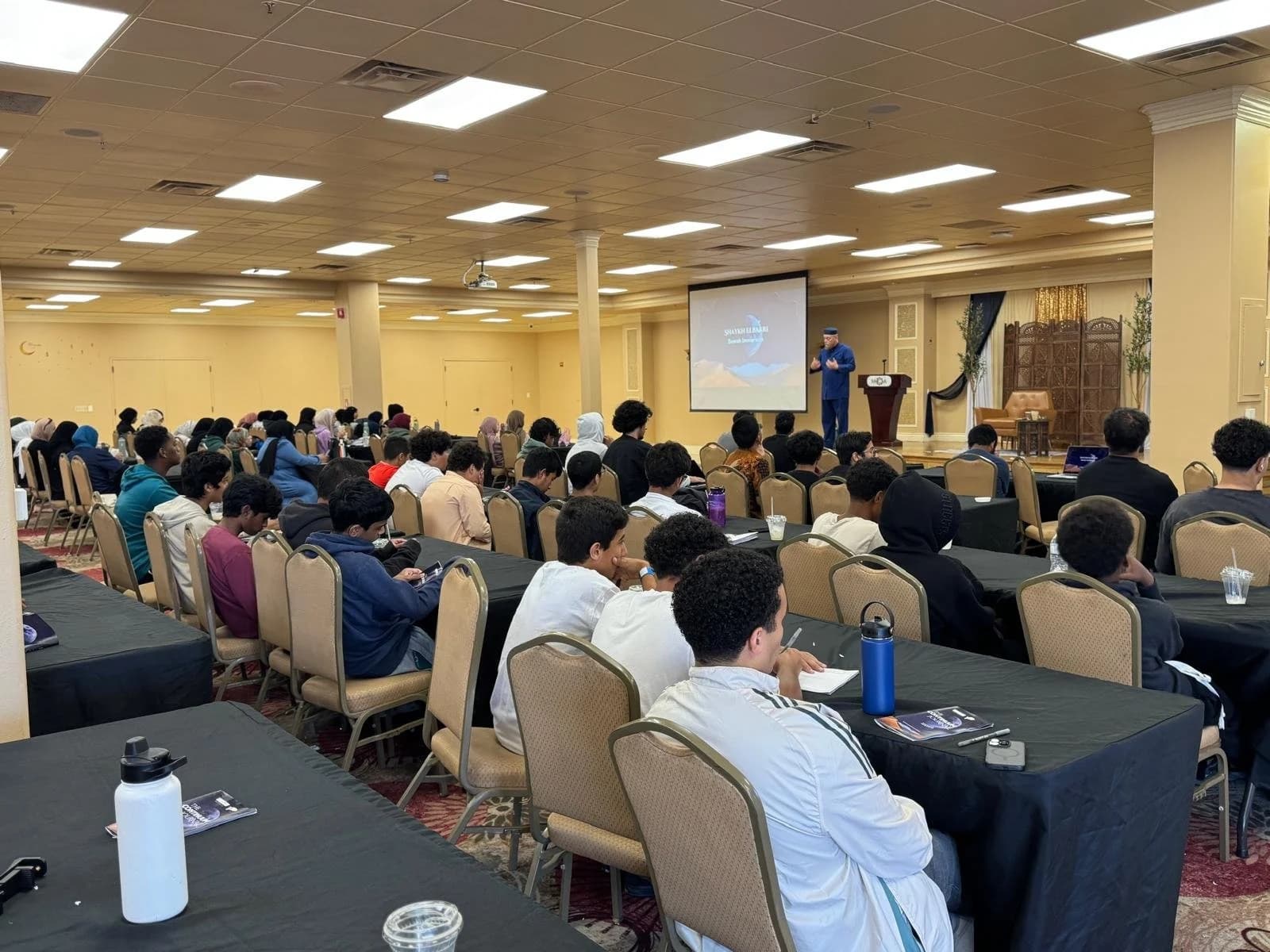 Teen attendees seated at long tables facing a speaker beside a projection screen in a conference hall.