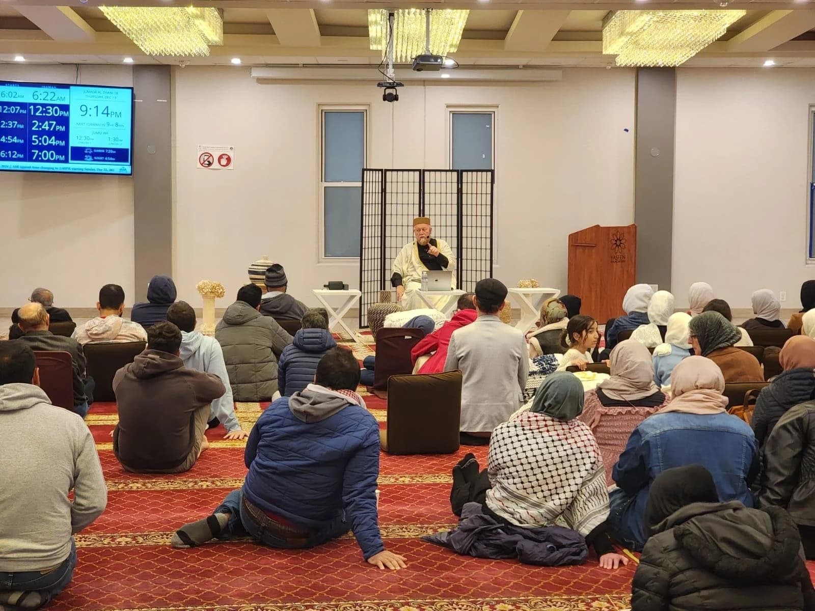 Seated teacher addressing attendees sitting on carpet in a small, bright prayer room with a folding screen behind him.